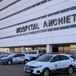 Fachada do Hospital Anchieta no DF com viaturas policiais, representando investigação de mortes suspeitas em UTI.