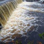 Reservatório do Descoberto transbordando, elevando risco de inundações no Distrito Federal, com água jorrando sobre a barragem e paisagem do Cerrado.