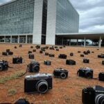 Vista de Brasília com Esplanada dos Ministérios e Congresso Nacional, destacando desafios urbanos e restrições orçamentárias, com câmera fotográfica simbolizando concurso.