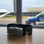 Tornozeleira eletrônica e dispositivo de bloqueio em delegacia no DF, com viatura policial ao fundo em Brasília.
