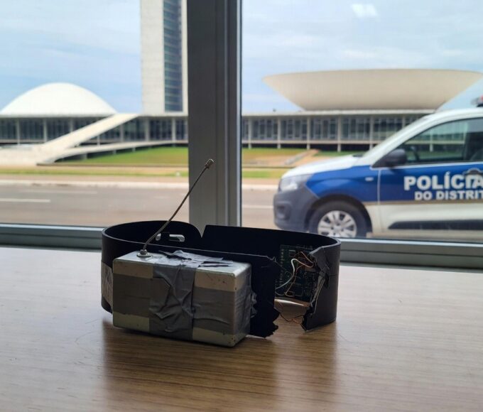 Tornozeleira eletrônica e dispositivo de bloqueio em delegacia no DF, com viatura policial ao fundo em Brasília.