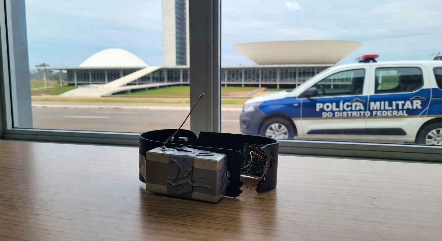 Tornozeleira eletrônica e dispositivo de bloqueio em delegacia no DF, com viatura policial ao fundo em Brasília.