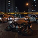 Cena de rua em Taguatinga, DF, com viaturas policiais e fita de isolamento após tiroteio, sem pessoas.