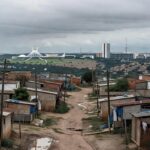 Vista panorâmica de Ceilândia com casas humildes e skyline de Brasília ao fundo, destacando desigualdades urbanas.