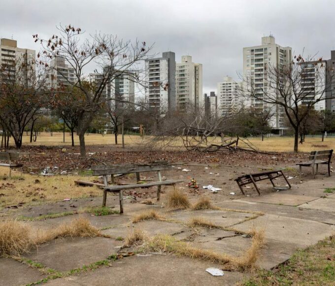 Parque abandonado em Águas Claras com grama seca, bancos quebrados e prédios ao fundo, destacando negligência em áreas verdes.